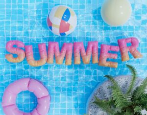Colorful inflatable letters spelling 'SUMMER' over a pool, with a multicolored ball and pink donut ring nearby.