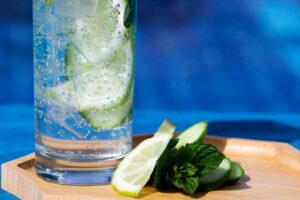 Tall glass of lime and cucumber sparkling water with ice, on a wooden tray by a blue pool, with lime wedges and mint garnish.