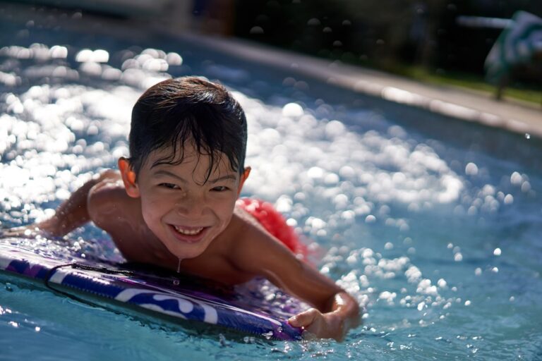 Smiling boy on a purple-and-white pool float, swimming in a sunny outdoor pool.