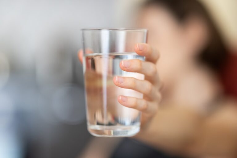 Close up of a hand holding a tall glass of water toward the camera with a blurred person in the background
