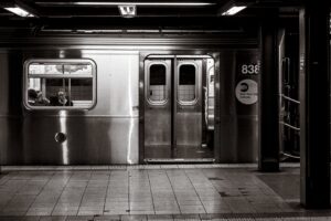 New York subway platform with a metal train car doors open and passengers visible through the window (black-and-white photo).