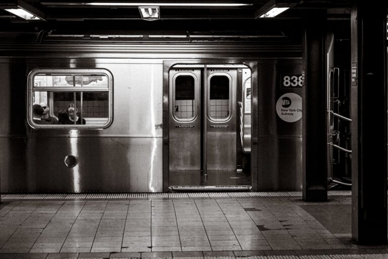 New York subway platform with a metal train car doors open and passengers visible through the window (black-and-white photo).