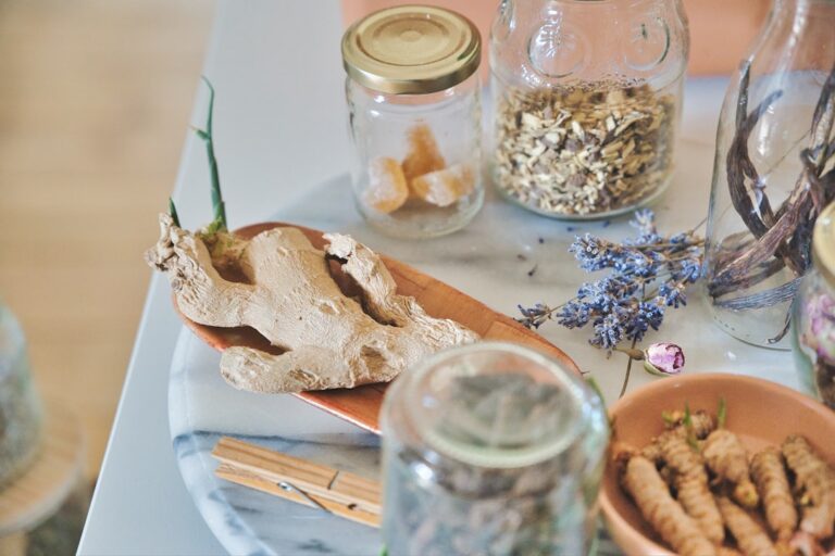 Ginger root on a wooden spoon atop a marble tray, with glass jars of dried herbs and brown sugar cubes in the background.