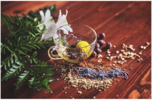 Clear glass teacup with dried lavender, chamomile, and other herbs on a wooden table beside fresh greens and white flowers.
