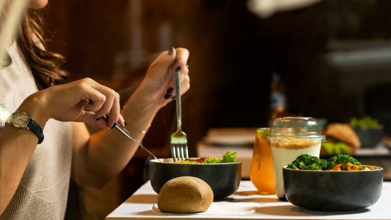 Howtoavoidfoodpoisoningnycstreetfood sickday Person dining at a table with bowls of salad and broccoli a bread roll and drinks nearby eating with a fork and knife