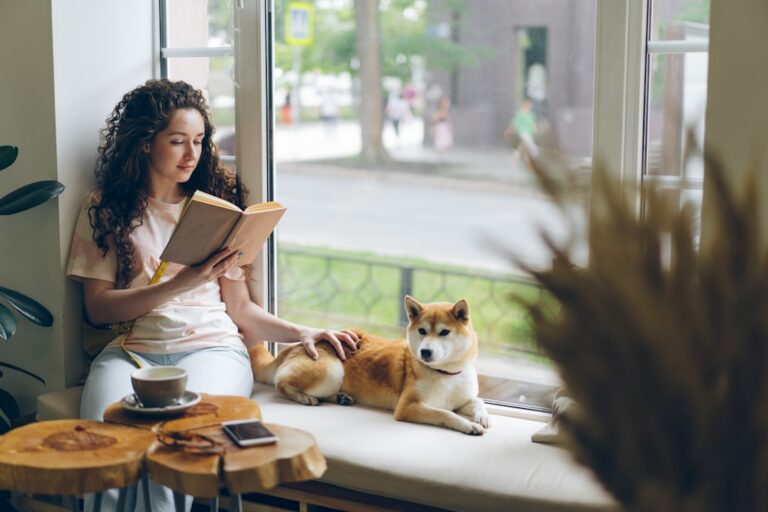 Woman reads a book by a sunny window, with a dog resting beside her on the sill.