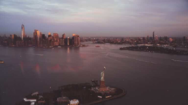 Aerial view of the Statue of Liberty on Liberty Island at dusk with the Manhattan skyline across New York Harbor behind it.