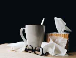 White mug with a straw beside a tissue box and scattered tissues, with glasses on a beige surface against a dark background.