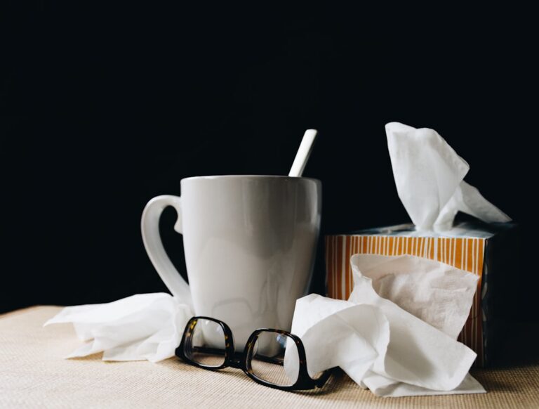 Pantrycoldremediesevidencebased sickday White mug with a straw beside a tissue box and scattered tissues with glasses on a beige surface against a dark background