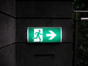 Blog 6 Green emergency exit sign with a running figure and a rightward arrow mounted on a dark wall