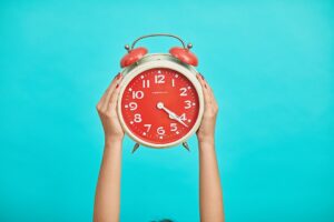 Red vintage alarm clock held up by two hands against a bright turquoise background, showing around 4:40.