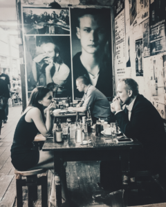Three people sit at a worn wooden table in a street-side cafe, talking over drinks with posters on the wall behind them.