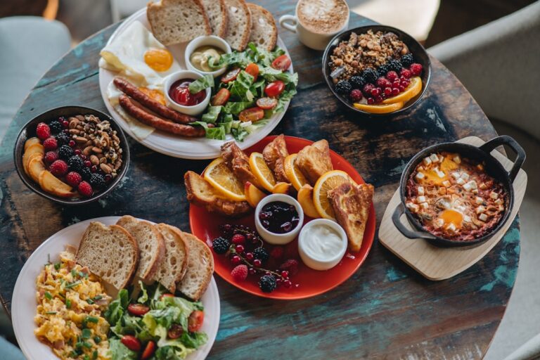 Top-down view of a rustic table with a breakfast spread: eggs, sausages, salad, bread, croissants, berries, yogurt, and a shakshuka skillet.