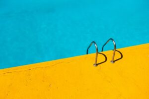 Blog 3 Metal pool ladder rails anchored to a bright yellow pool edge with blue water in the background.